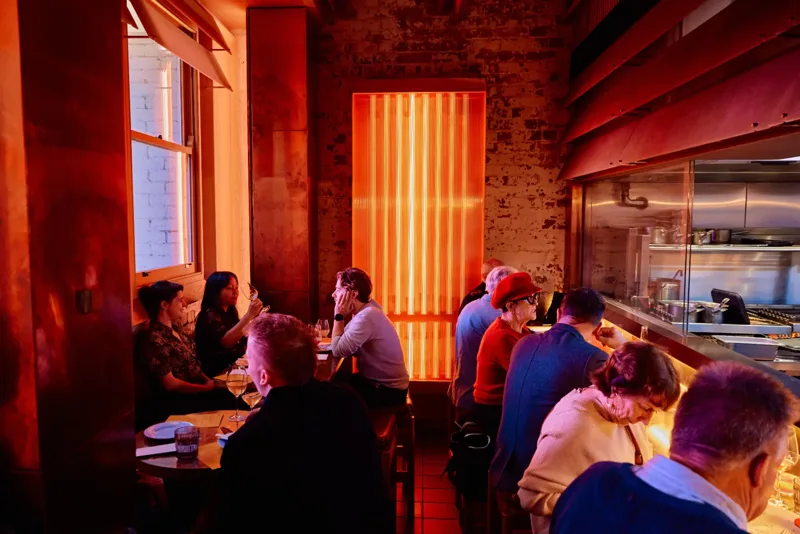 Diners at tables and the kitchen pass in the warm red-lit dining room at BKK