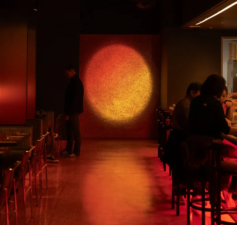 Dimly lit dining room at Sachi with a glowing sunset mural and guests seated along the counter